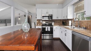 A modern kitchen with white cabinets, stainless steel appliances, a wooden table, and a window with blinds. - Family Favorite_Sleeps 8_Roomy_Game Room (Lenexa)