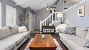 A living room with a wooden coffee table, two grey sofas, a staircase, and a white chair. - Family Favorite_Sleeps 8_Roomy_Game Room (Lenexa)