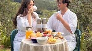 Um casal sentado à mesa apreciando o café da manhã com diversas comidas e bebidas, com vista para montanhas.
