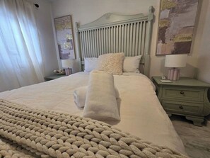 A neatly made bed with a knitted blanket, a headboard, a bedside table with a lamp, and a nightstand with a drawer.