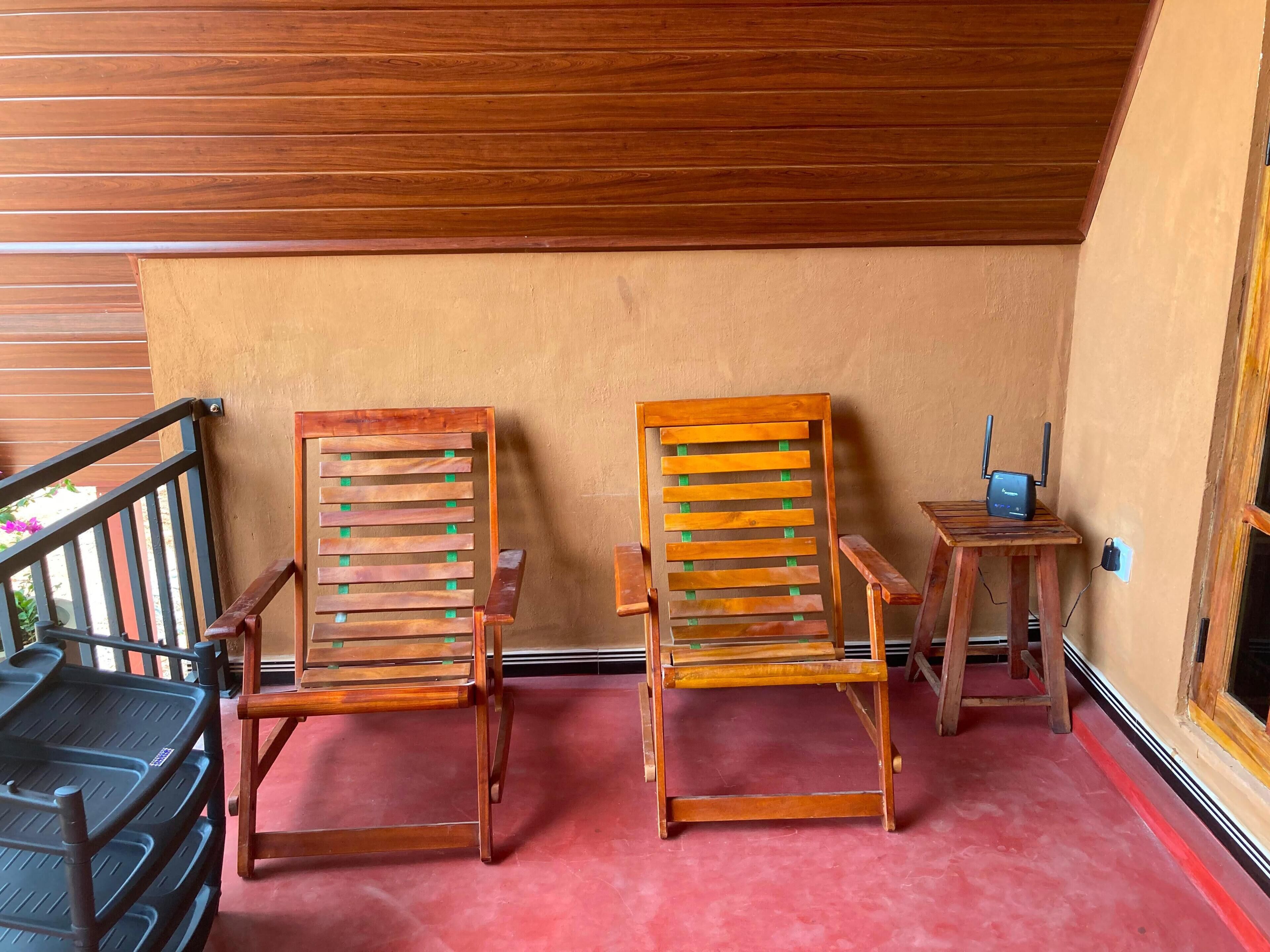 Dos sillas de madera con respaldos de listones y una pequeña mesa de madera con una computadora portátil encima, frente a una pared color beige y un techo de madera.