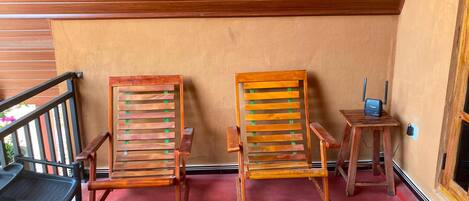 Dos sillas de madera con respaldos de listones y una pequeña mesa de madera con una computadora portátil encima, frente a una pared color beige y un techo de madera.