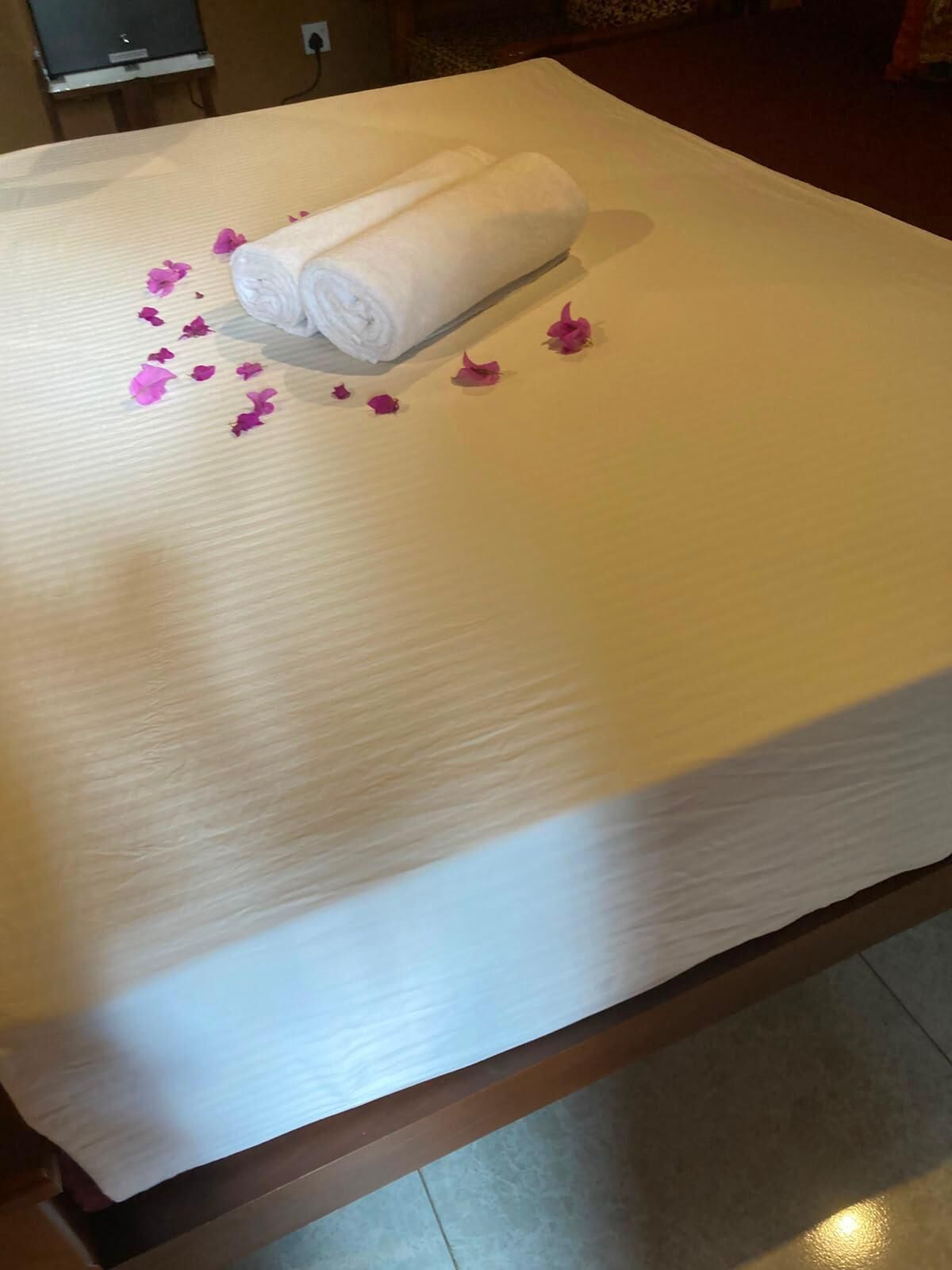 Una cama bien hecha con una toalla blanca enrollada y pétalos de flores rosas esparcidos.