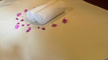 Una cama bien hecha con una toalla blanca enrollada y pétalos de flores rosas esparcidos.