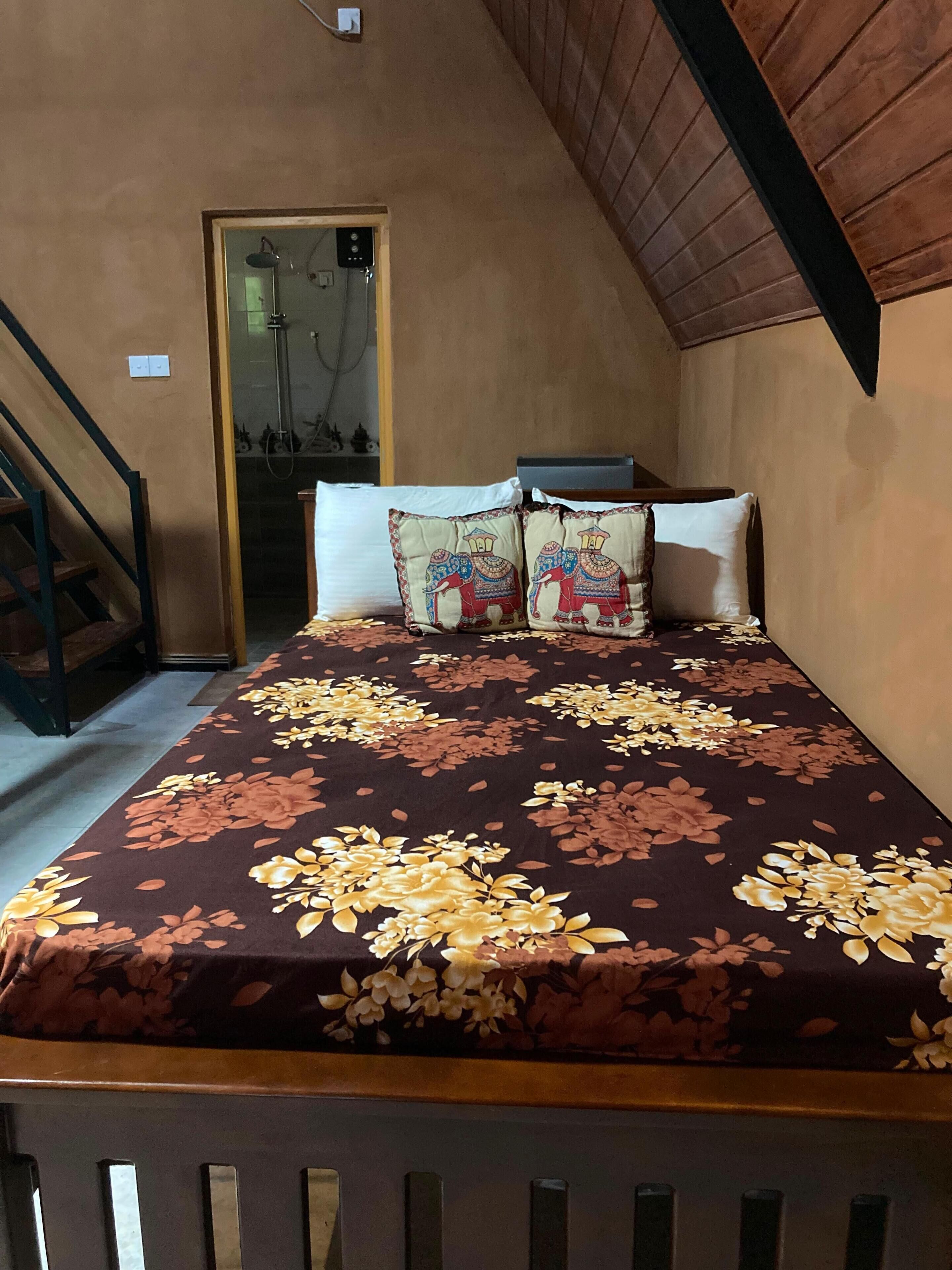 Une chambre à coucher avec un lit, deux oreillers ornés de motifs d’éléphants, une tête de lit en bois, un escalier et une porte donnant sur une salle de bain.
