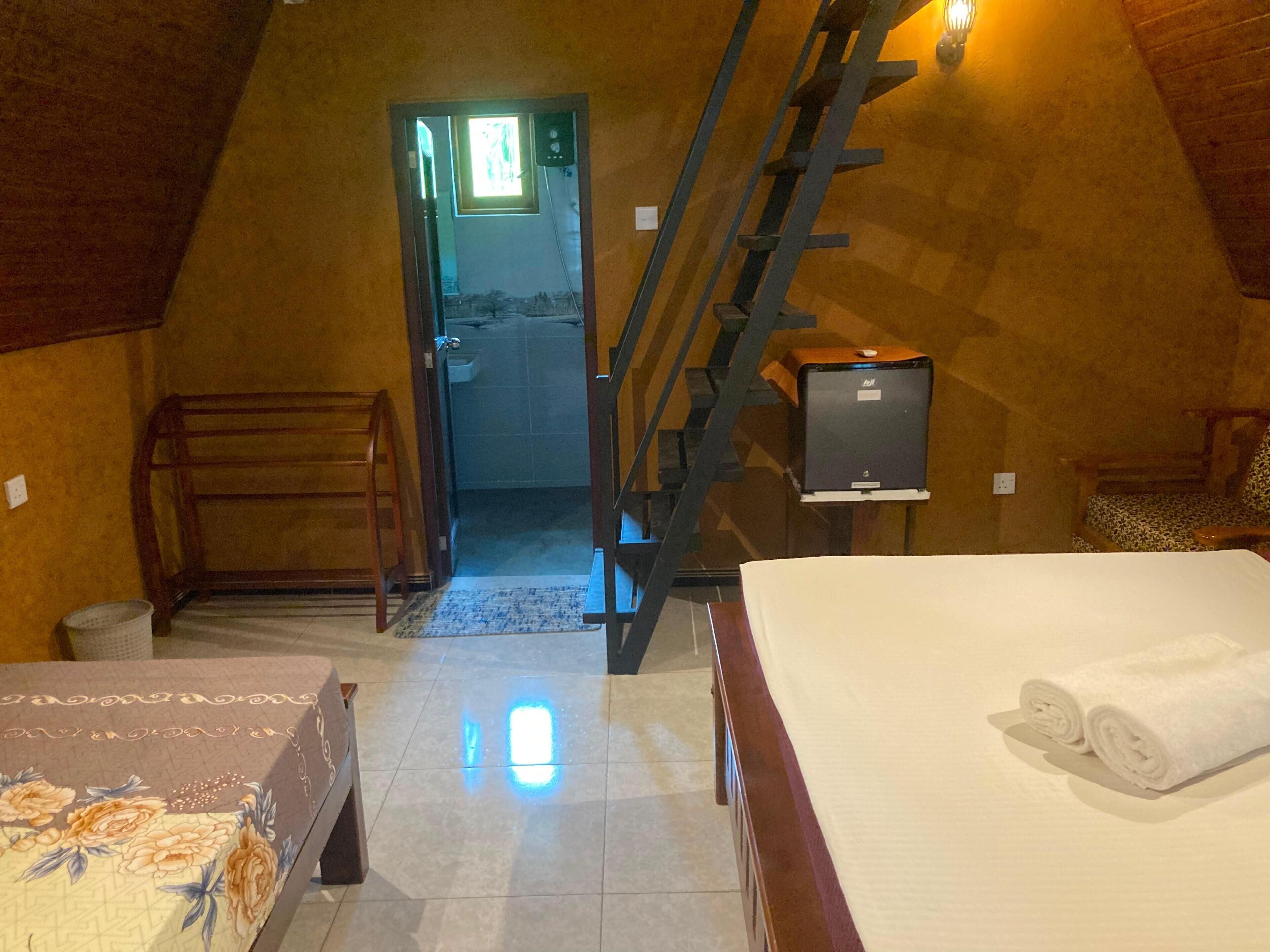 A bedroom with a bed, a chair, a desk, a ladder, and a bathroom visible through an open door.