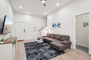 Ein modernes Wohnzimmer mit einer Eckcouch, einem Flachbildfernseher an der Wand, einem kleinen runden Tisch und einer Lampe.