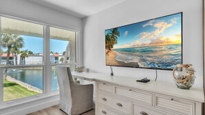 A modern room with a large window overlooking a pool and beach, a flat-screen TV on a white dresser, and a decorative vase. - Runaway Bay 276 by Duncan Real Estate (Bradenton Beach)