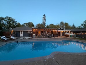 A swimming pool with lounge chairs, a covered patio, and a building with string lights. - Private Resort • Pool, Spa + Sauna (Grants Pass)