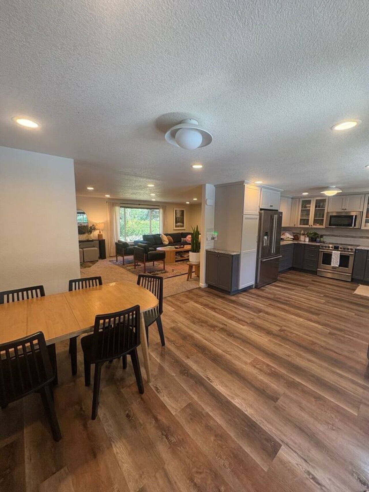 A spacious living area with a dining table, chairs, a kitchen with modern appliances, and a large window providing natural light.