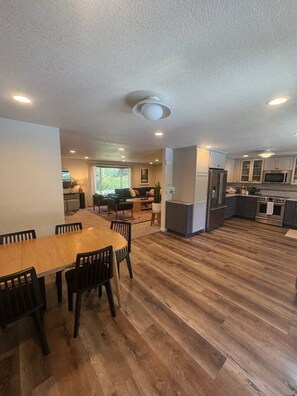 A spacious living area with a dining table, chairs, a kitchen with modern appliances, and a large window providing natural light. - Private Resort • Pool, Spa + Sauna (Grants Pass)