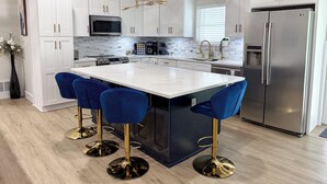 A modern kitchen with white cabinets, a central island with blue bar stools, stainless steel appliances, and a wooden ceiling beam. - Woodland Retreat/Pool+Patio+Firepit+SunsetViews (Weatherford)
