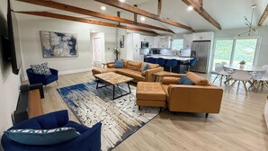 A modern living room with leather sofas, a coffee table, and blue chairs. A kitchen with white cabinets and a dining area with a white table and chairs is visible. - Woodland Retreat/Pool+Patio+Firepit+SunsetViews (Weatherford)