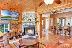 A cozy cabin interior with a stone fireplace, wooden walls, a television, and a dining area.
