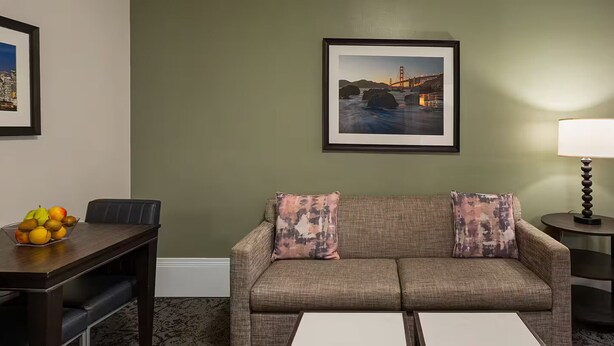 Ein Wohnzimmer mit einer Couch, einem kleinen Tisch mit Obst, einer Lampe und einem gerahmten Bild an der Wand.
