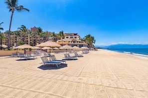 Ein Strand mit Liegestühlen und Sonnenschirmen, im Hintergrund ein Hotelgebäude und ein klarer, blauer Himmel.