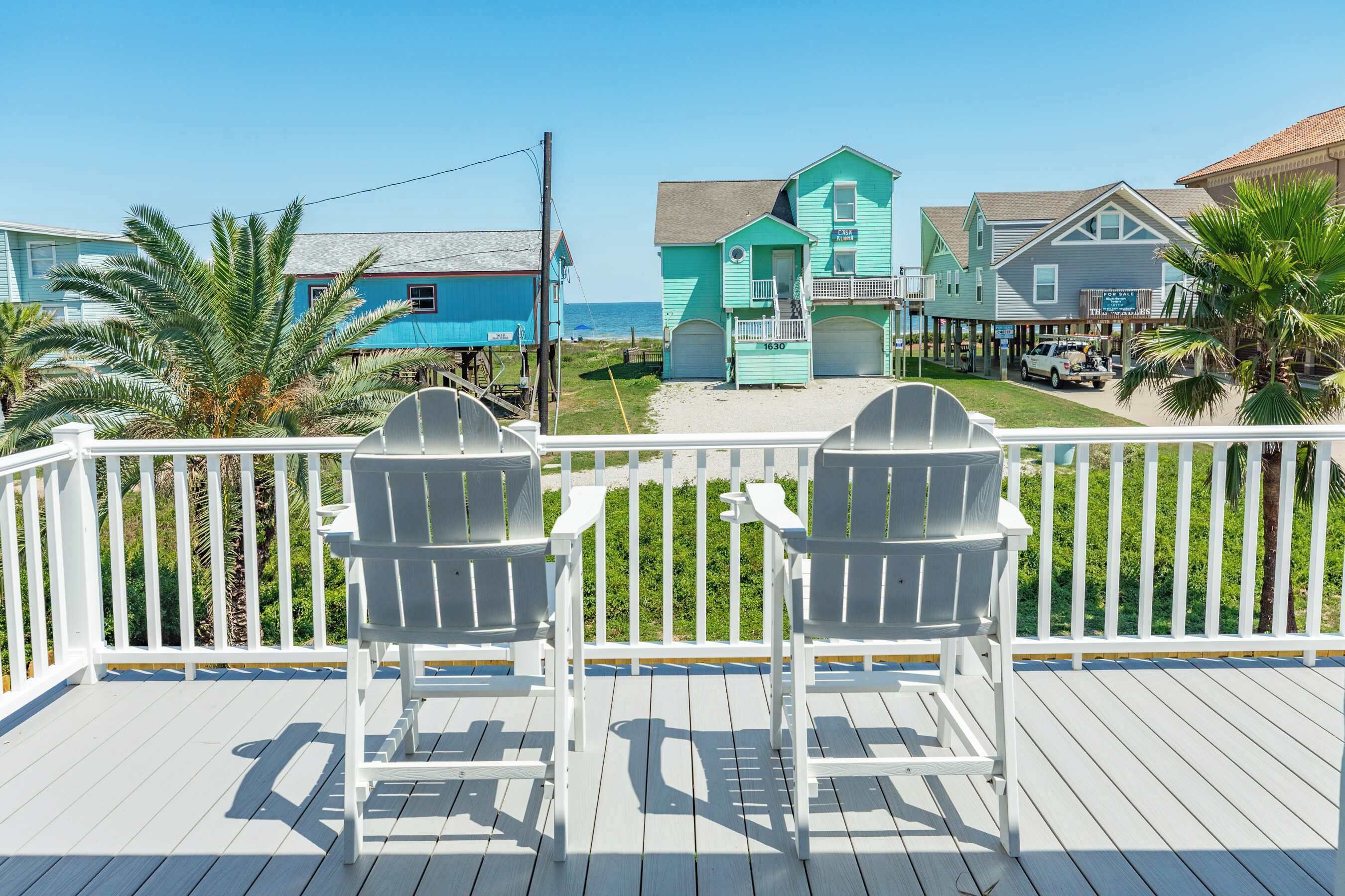 Una terraza con sillas Adirondack, vista a un edificio turquesa y una playa al fondo.