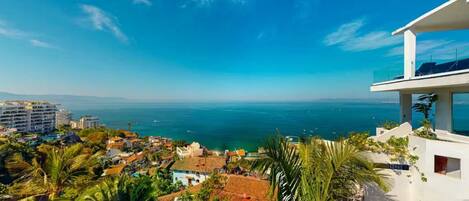 A coastal view with a balcony overlooking the sea, buildings in the background, and lush greenery.