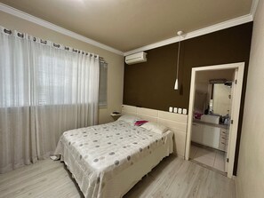 A bedroom with a bed, curtains, an air conditioning unit, a bedside table, and a bathroom visible through an open door. - Complete and cozy house for 7 people. (Maringá)