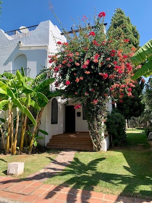 Ein weißes Gebäude mit einer kleinen Veranda, einer Treppe zum Eingang und einem Garten mit Bananenpflanzen und blühenden Sträuchern.