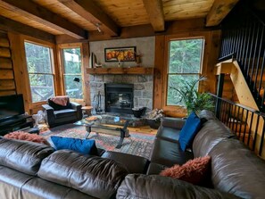 Ein Blockhaus-Interieur mit Kamin aus Stein, Ledersofas, einem Fernseher und großen Fenstern.