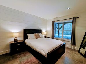 A bedroom with a large bed, two bedside tables, a window with curtains, and a view of the outdoors.