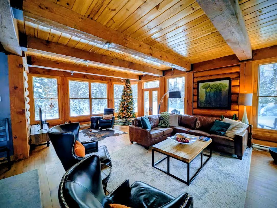 L’intérieur d’une cabane en rondins, avec un plafond en bois, de grandes fenêtres, une cheminée, des meubles en cuir et un sapin de Noël.