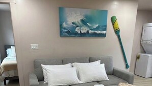 Un sofá gris con cojines blancos, un cuadro azul, una escoba verde y una lavadora blanca.