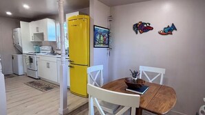 Una cocina compacta con armarios blancos, un refrigerador amarillo y una zona de comedor con una mesa de madera y sillas blancas.
