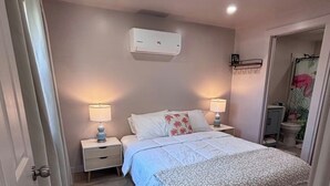 Un dormitorio con cama, dos mesitas de noche, aire acondicionado de pared y una puerta que da a otra habitación.