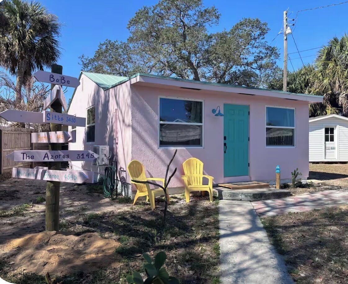 A small pink building with a green door, two yellow chairs outside, and directional signs.