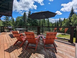 Eine Holzterrasse mit roten Stühlen und einem Tisch, einem großen Sonnenschirm und Blick auf Bäume und ein Gebäude.