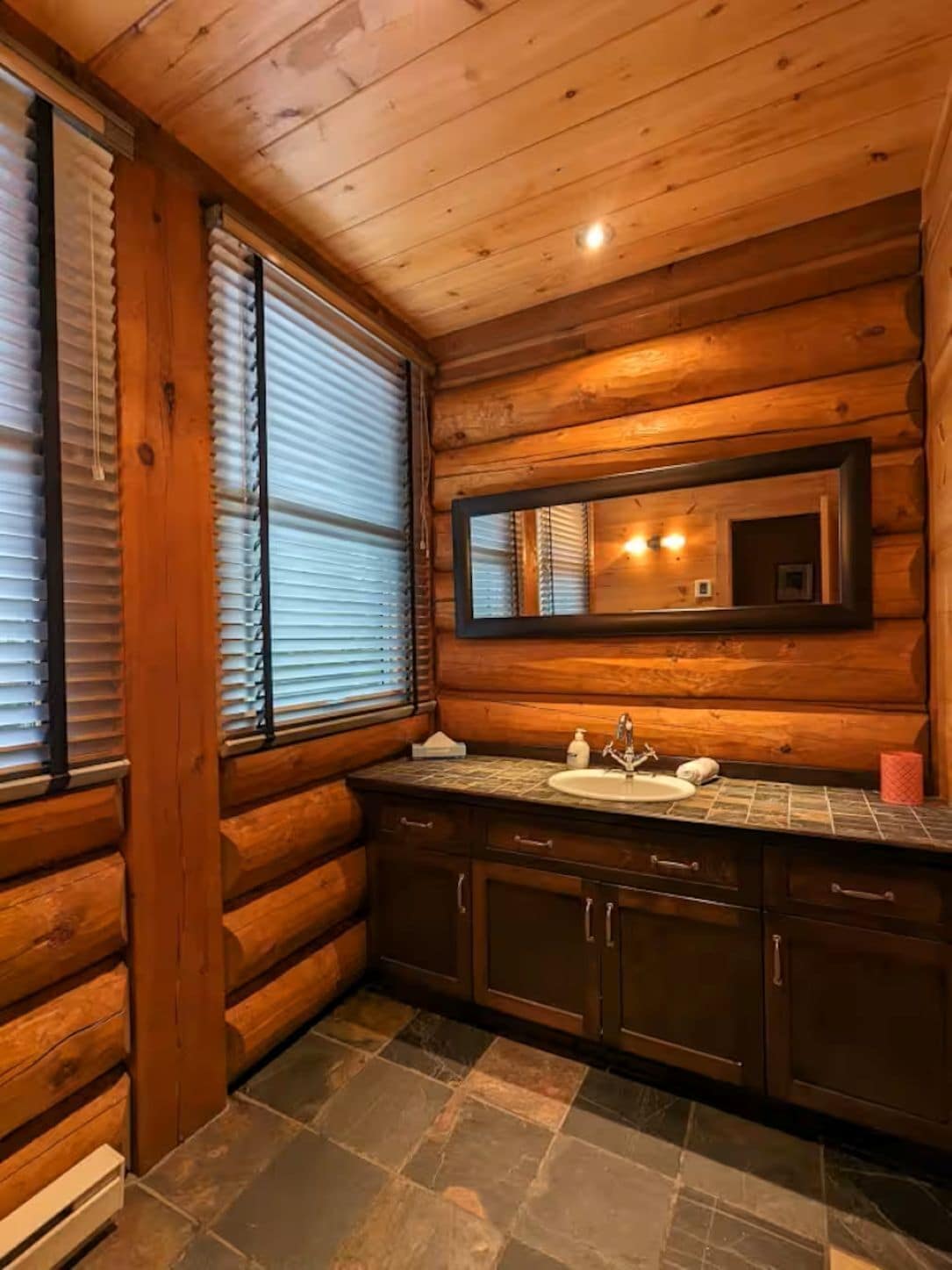 A log cabin bathroom with a stone-tiled floor, wooden walls, a large mirror, and a sink with a faucet.