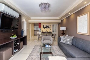 Een moderne woonkamer met een flatscreen televisie, een glazen salontafel, een grijze bank en een eethoek op de achtergrond.