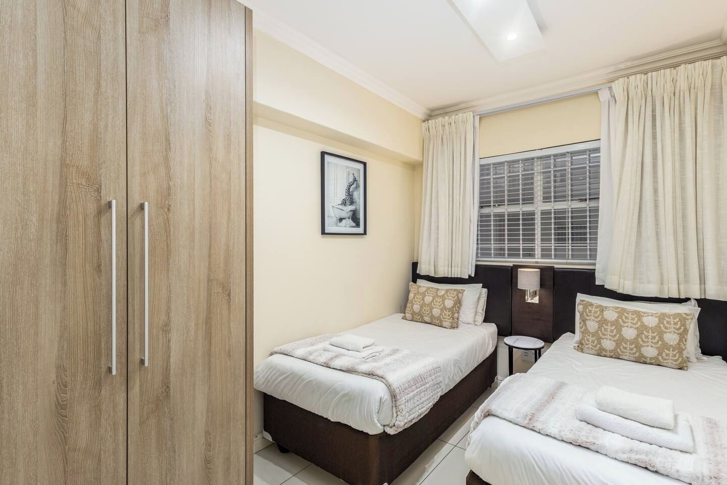A hotel room with two beds, a wardrobe, a bedside table, a window with curtains, and a wall-mounted picture.