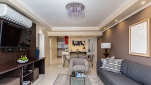 A modern living room with a flat-screen TV, a glass coffee table, a grey sofa, and a dining area in the background. - New Cumberland 405 by CTHA (Cape Town)