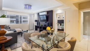 A modern living room with a glass dining table, comfortable seating, a flat-screen TV, and a large window. - New Cumberland 405 by CTHA (Cape Town)