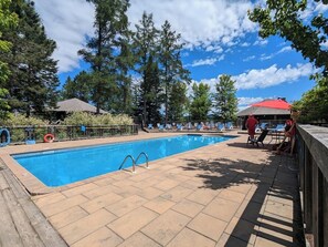 A rectangular swimming pool with a tiled deck, surrounded by trees and a clear sky.