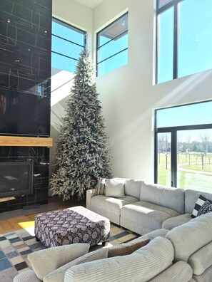 A modern living room with a large window, a fireplace, a sofa, and a Christmas tree. - Forest Edge Luxury  Estate (Puslinch)