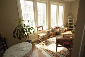 Ein Zimmer mit großen Fenstern, einem Holzstuhl, einem Korbssessel, einem gemusterten Teppich und einer weißen Kommode.