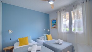 Ein Zimmer mit zwei Betten, jeweils mit blauer Bettdecke und gelbem Kissen, einem kleinen Holztisch, einem Deckenventilator und einem Vorhangfenster.