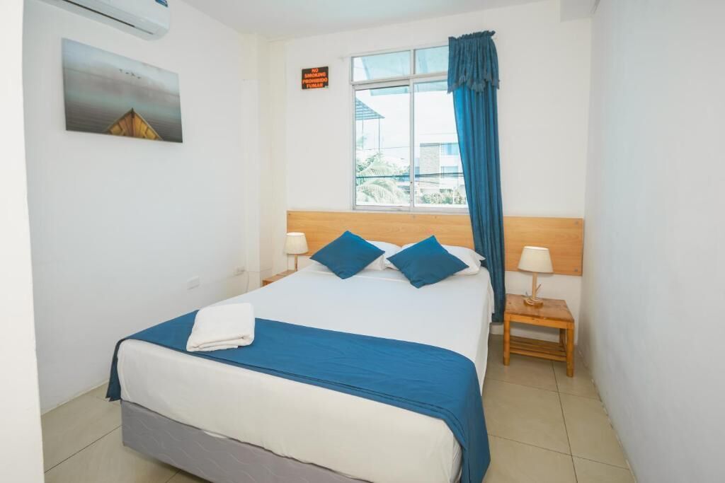 A bedroom with a bed, two bedside tables, a window with curtains, a wall-mounted air conditioner, and a framed picture.