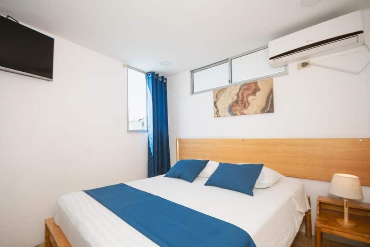 A bedroom with a bed, a wall-mounted air conditioner, a window with blue curtains, and a painting.