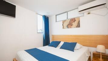 A bedroom with a bed, a wall-mounted air conditioner, a window with blue curtains, and a painting.