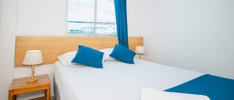 Habitación de hotel con cama, mesita de noche con lámpara, ventana con cortinas azules y vista a edificios.
