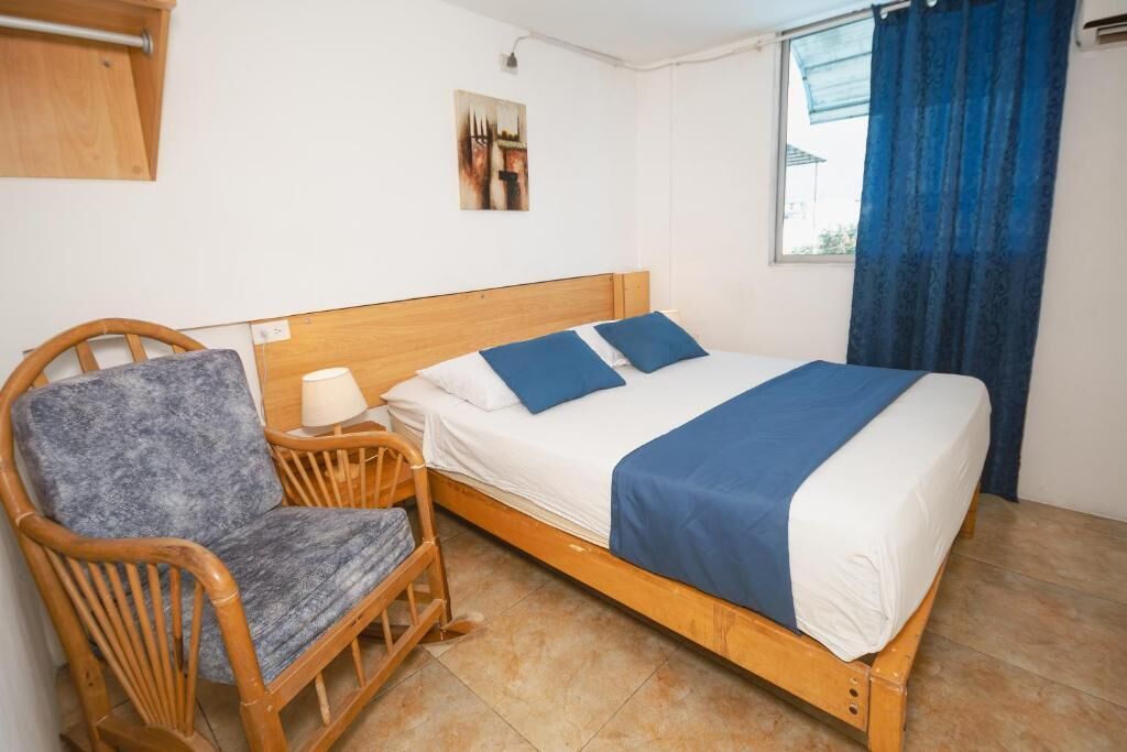 A single bed with a wooden frame, a chair with a cushion, a wall-mounted lamp, and a window with blue curtains.