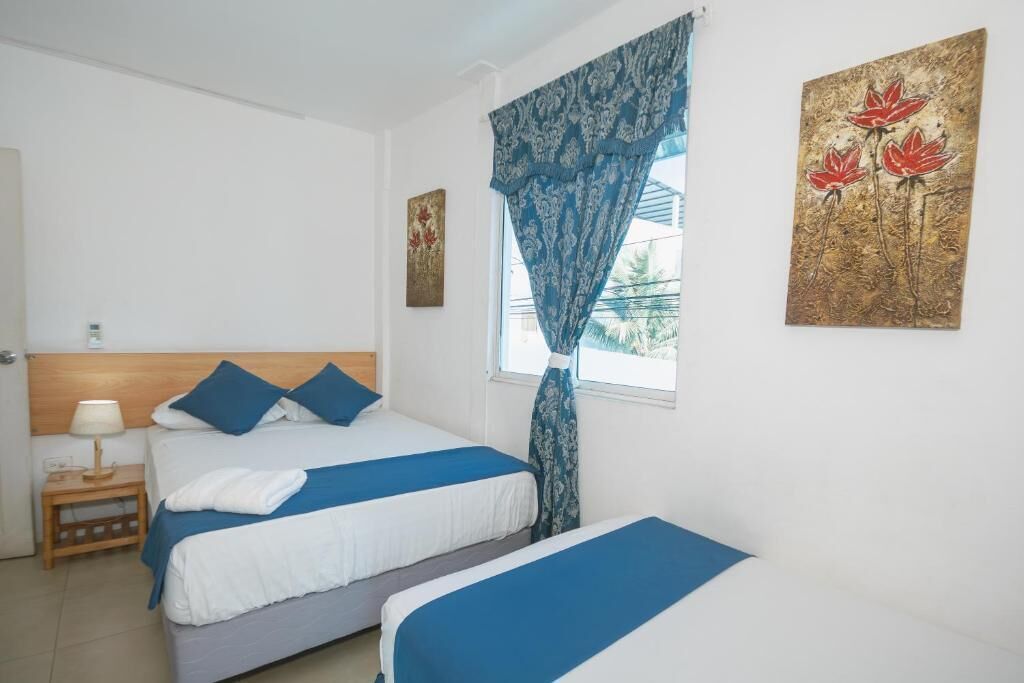 Ein Hotelzimmer mit zwei Betten, einem Nachttisch, einem Fenster mit gemusterten Vorhängen und Bildern an den Wänden.