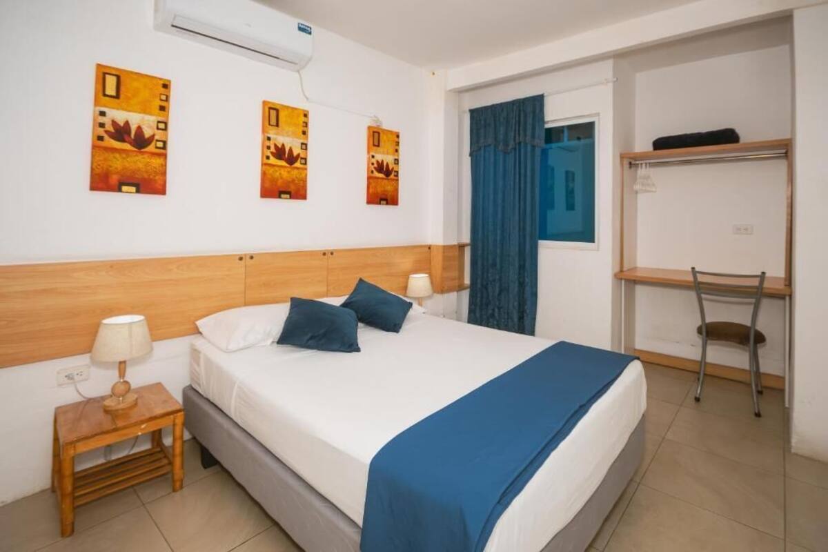 A hotel room with a bed, bedside tables, a desk, and artwork on the wall.