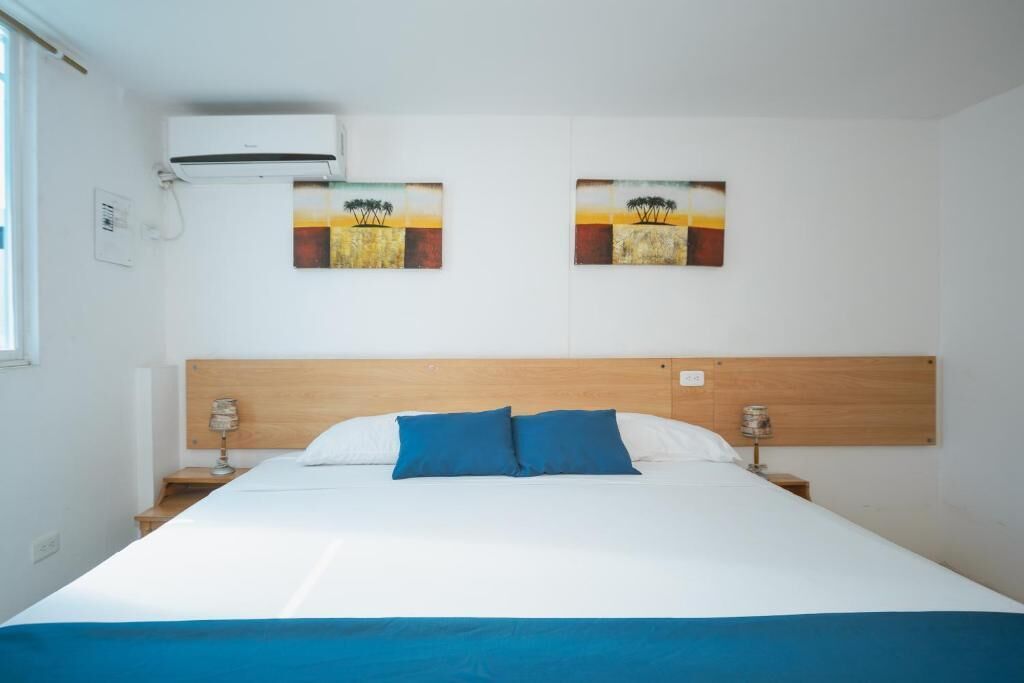 Habitación de hotel con una cama, dos cuadros en la pared, cabecera de madera, mesitas de noche con lámparas y un equipo de aire acondicionado.