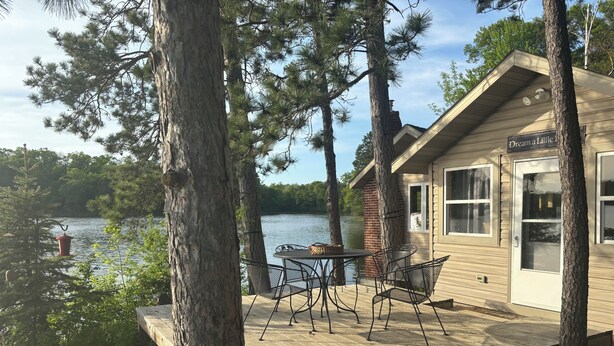 A cabin with a deck, a table, and chairs, overlooking a lake.